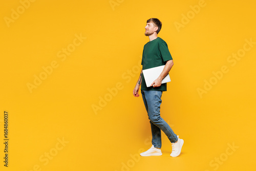 Fotografie Full body side profile view young IT man wear green t-shirt casual clothes hold closed laptop pc computer walking going isolated on plain yellow orange background studio portrait