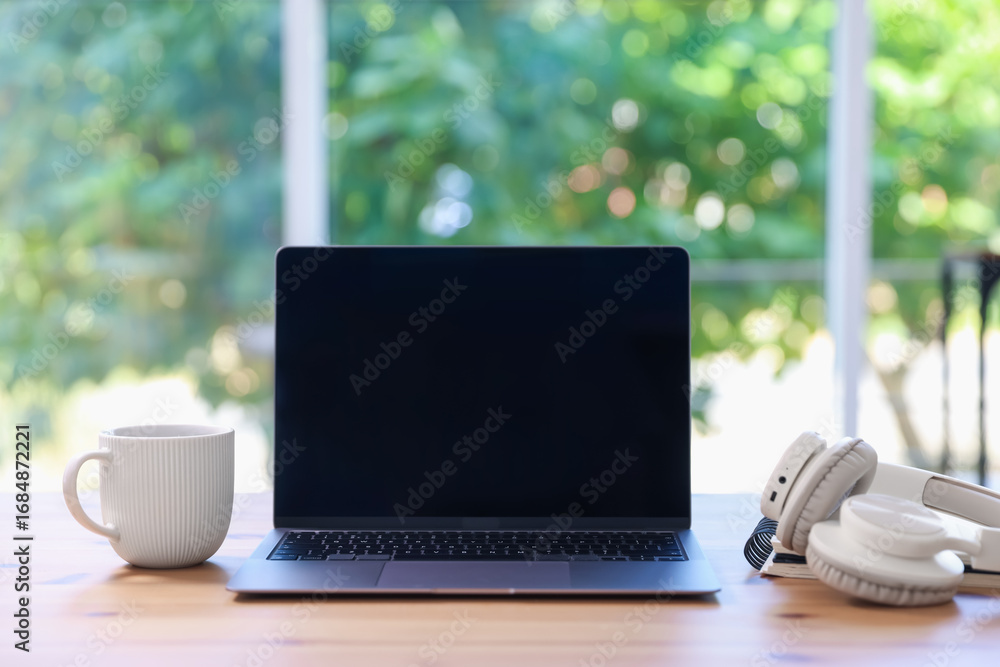 Fototapeta premium Home workspace. Laptop, cup of drink and headphones on wooden desk near window indoors