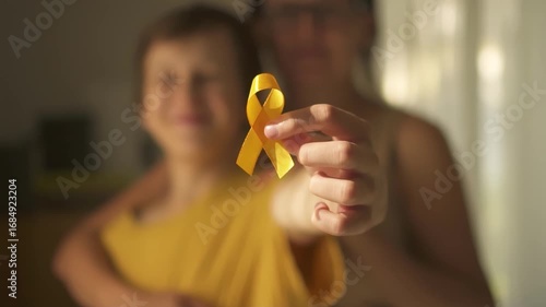Teen boy holds yellow awareness ribbon while mother embraces him from behind, showing family support for pediatric cancer awareness.