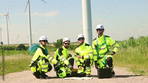 Team technical engineers in uniforms armed with blueprints and toolboxes sits with determination on the field wind turbine power plant site working as a team plan and monitor maintenance schedules.
