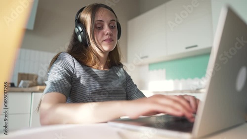 Working on laptop with headphone reaching hand toward viewer while woman types on laptop keyboard at kitchen desk wearing watch and headset showing online work and gesture during remote work session