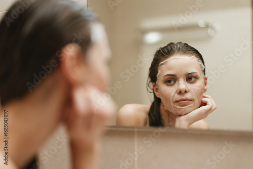 Free Download Pensive young woman with facial mask, gazing thoughtfully in bathroom mirror. Skincare and self-care routine concept. Relaxation and beauty routine. - GFXLoad