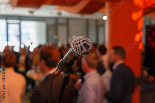 Slika na platnu A microphone stands in focus as people mingle in a lively setting, preparing for an engaging event