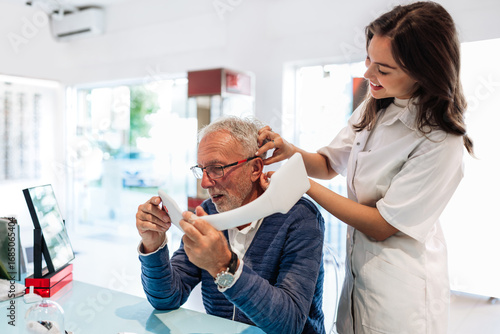 Audiologist is helping a senior man put on a hearing aid. While he looks in a mirror to see the process in a modern clinic setting. Showcasing the importance of hearing healthcare for the elderly