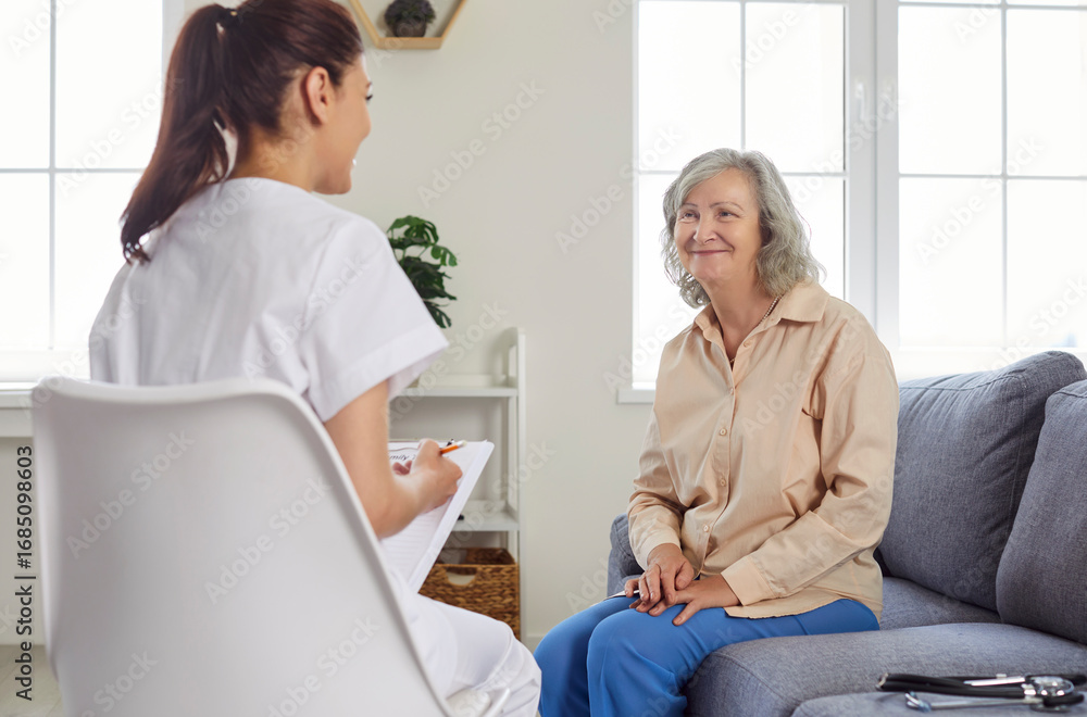 Obraz premium Friendly nurse or doctor visits senior patient at home. Young medical worker in uniform with clipboard sitting on chair and talking to happy smiling old woman on couch. Geriatrics, health care concept