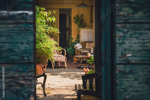 Rustic Italian patio with vintage furniture, plants and cozy atmosphere, captured through open wooden doors. Warm light, intimate mood, Mediterranean style.