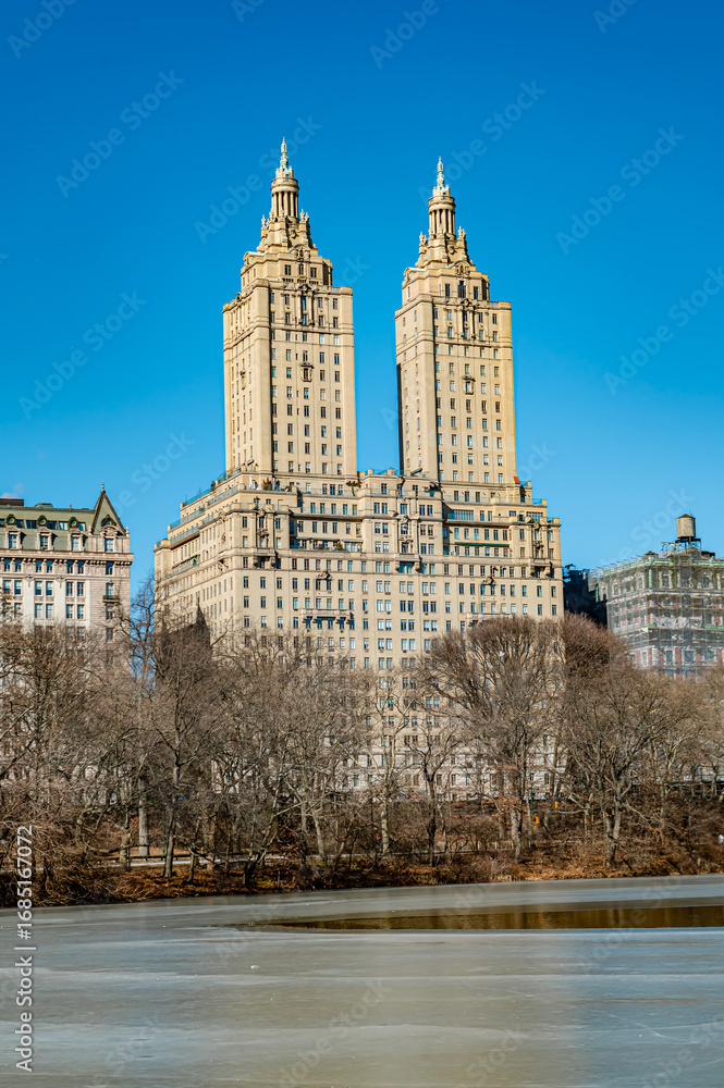 Obraz premium Central Park view of iconic twin towers. Twin towers rise over Central Park, framed by bare trees under a clear blue sky in New York City.