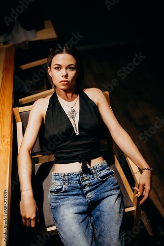 Moody portrait of a young woman reclining on a chair under low, cinematic light. Black halter top, denim, tattoo and jewelry visible. Warm highlights and deep shadows shape a calm yet confident vibe.