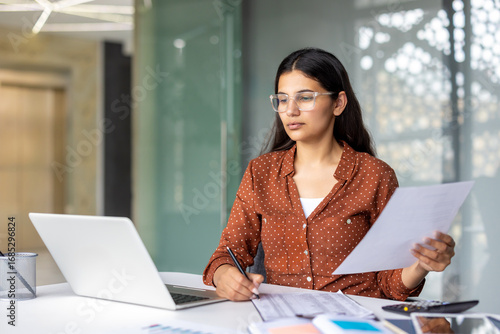 Tapet Serious and pensive business woman behind paper work inside office, female financier worker thinks about contracts and reports with charts and graphs, successful woman uses laptop at work