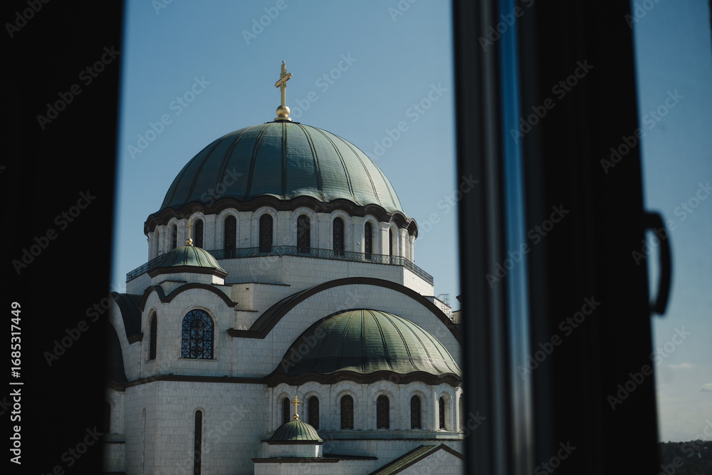 Fototapeta premium View of Saint Sava Temple in Belgrade framed by a window, with its green domes and golden cross shining under the blue sky