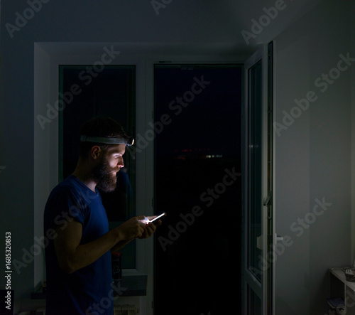 Man with beard using smartphone in dark room, wearing headlamp for light during night or emergency