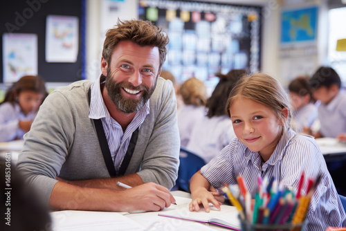 smiling male primary school teacher helping a student with writing at a clean desk, colorful but uncluttered classroom background