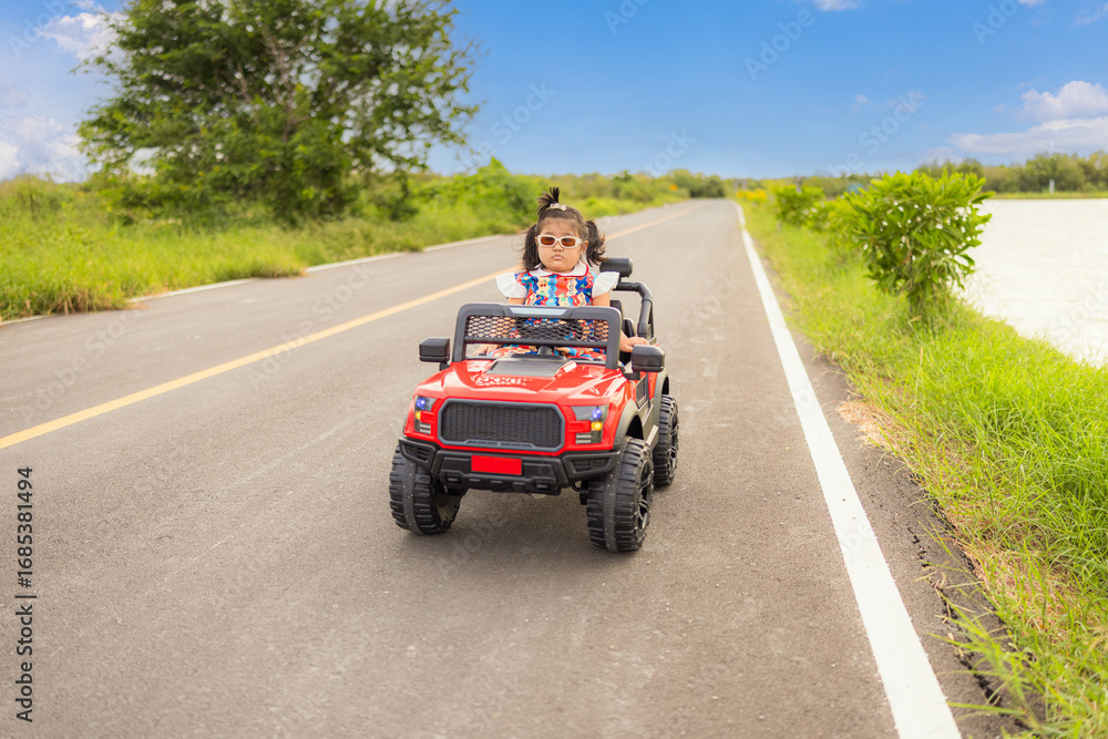 Fototapeta premium A cute little Asian girl is happily riding her toy car on a country road during her summer vacation,A little girl rides a toy car on a concrete road.