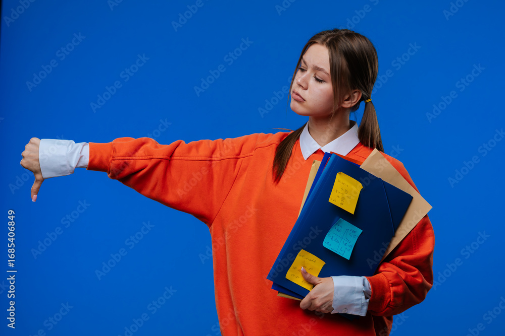 Naklejka premium A young female student gives a thumbs down gesture while holding folders and notes in her hands.
