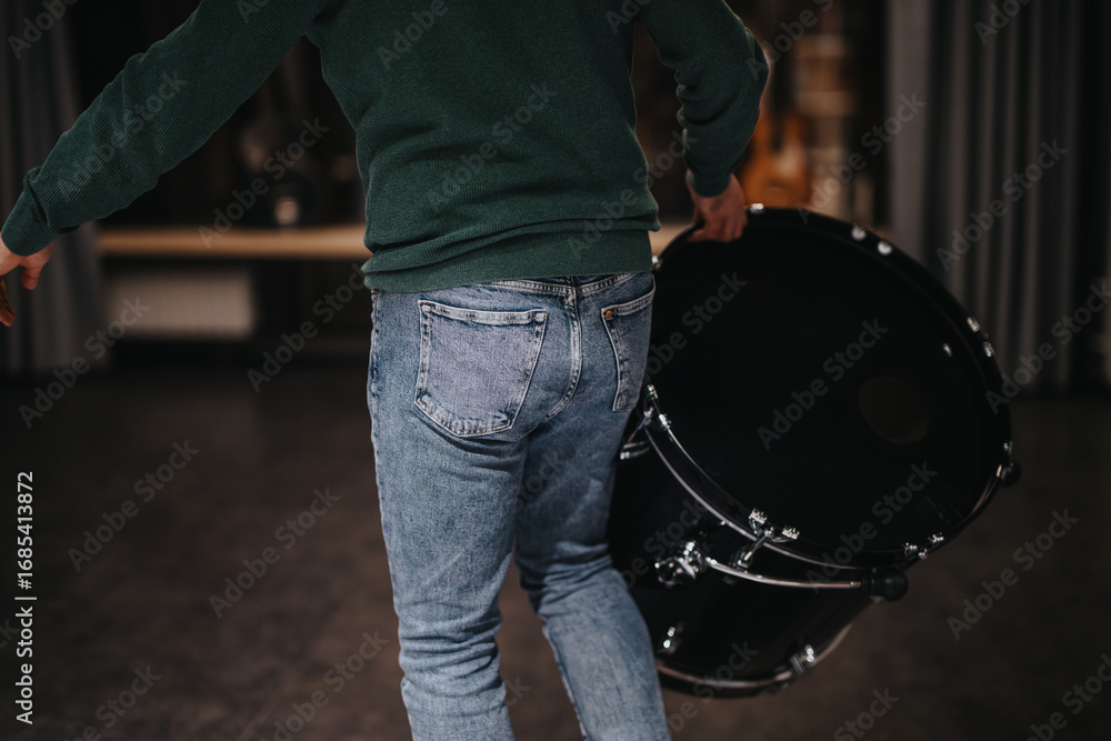Obraz premium A person dressed casually in a sweater and jeans carries a large drum in a dimly lit music studio. The scene suggests preparation or practice, capturing a behind-the-scenes moment in music production.