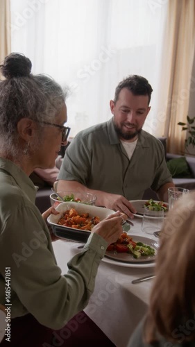 Vertical view of senior woman filling plate with sweet potatoes and passing ceramic dish to son-in-law during festive lunch at home