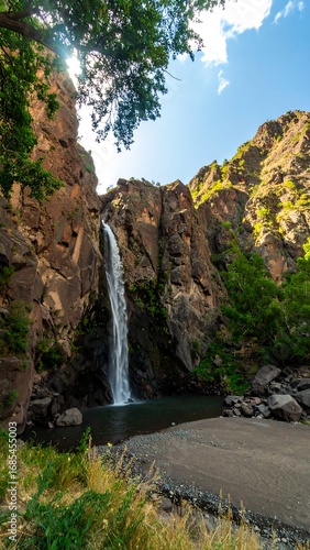 Waterfall cascading down rocky cliffs, surrounded by greenery and sunlight
