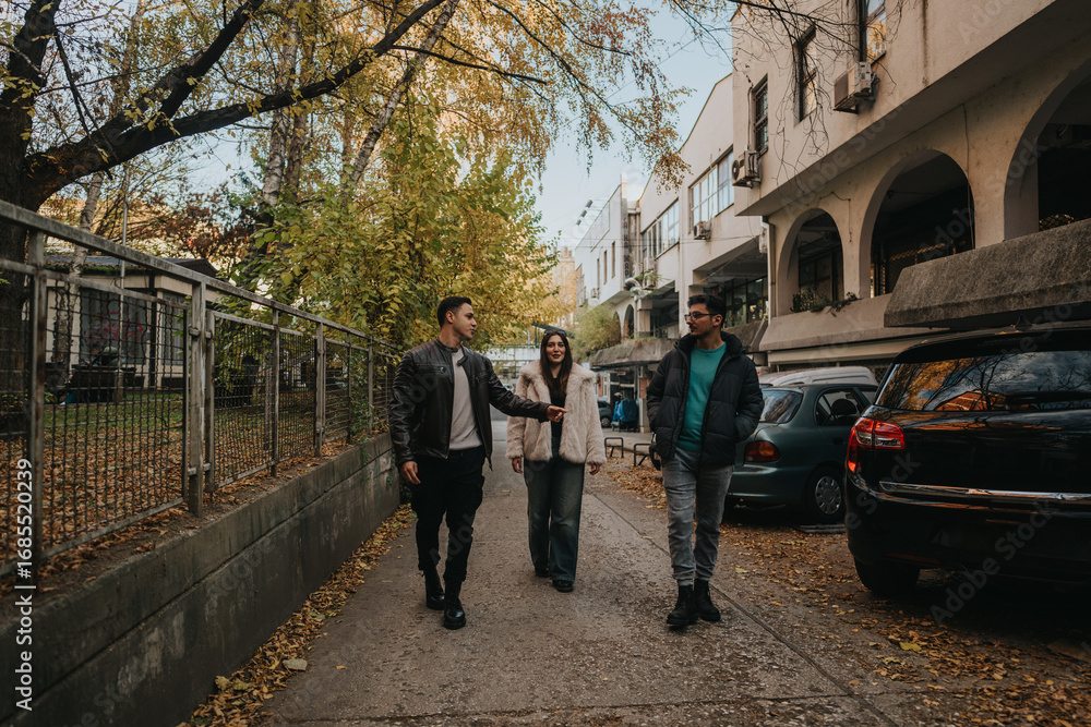 Obraz premium Three friends walking together through a city street during autumn, surrounded by falling leaves.