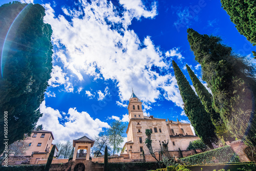 Historic Partal Palace tower at Alhambra complex in Granada Spain against dramatic blue sky with white clouds and Mediterranean cypress trees framing the view.
