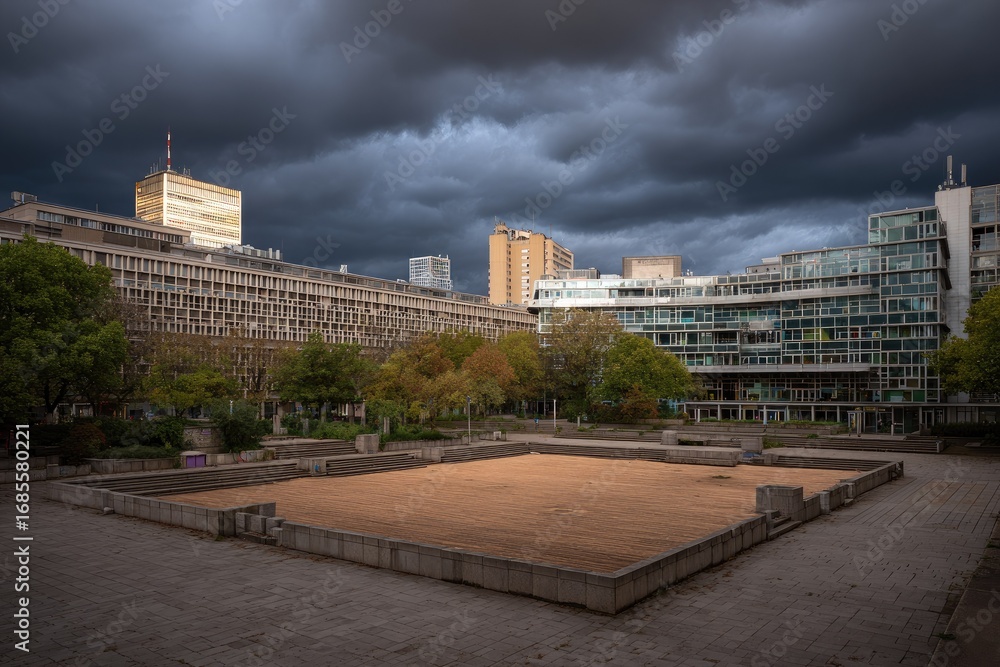 Fototapeta premium Empty square courtyard surrounded by modern buildings under a dramatic sky.