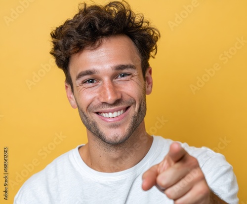 A cheerful man with a warm smile and curly hair directs his finger towards the viewer against a vibrant yellow backdrop.