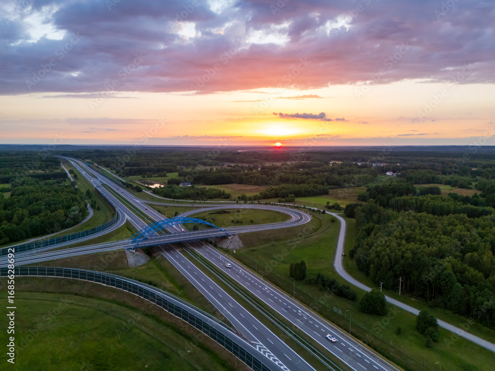 Fototapeta premium Aerial view of a highway intersection surrounded by lush green fields at sunset. Roads lead into the horizon, showcasing the beauty of nature and infrastructure.