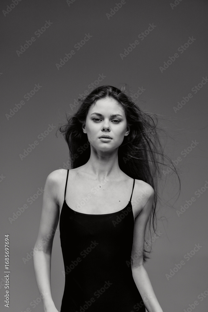 Fototapeta premium Black and white portrait of a serious young woman with long hair wearing a black dress against dark background