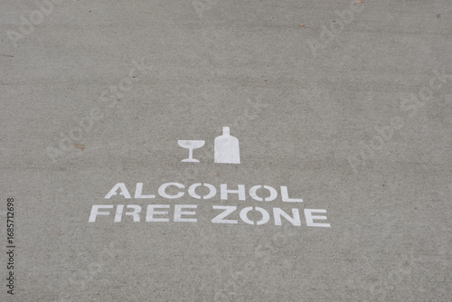 A sign for alcohol free zone on the street or asphalt road written in Nelson, New Zealand with a picture of glass and bottles. The sign is painted in white color.
