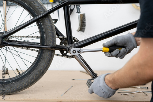 A person in gloves is tightening a bicycle crank on a bike frame using a ratchet wrench in a well-equipped workshop with tools in the background.