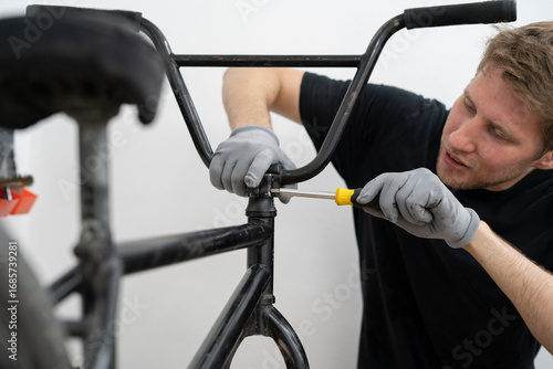 A person is carefully installing new bicycle handlebars in a workshop, using a screwdriver while wearing gloves. The bike rests on a stand for easy access.