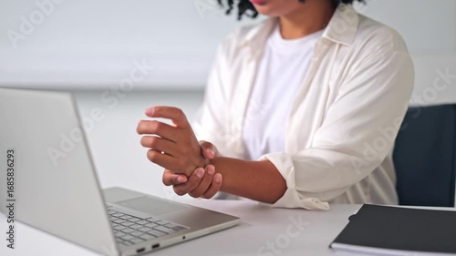 Professional businesswoman in a white shirt experiences wrist discomfort while working on a laptop in a bright office. She massages her painful wrist, expressing strain from prolonged computer use.