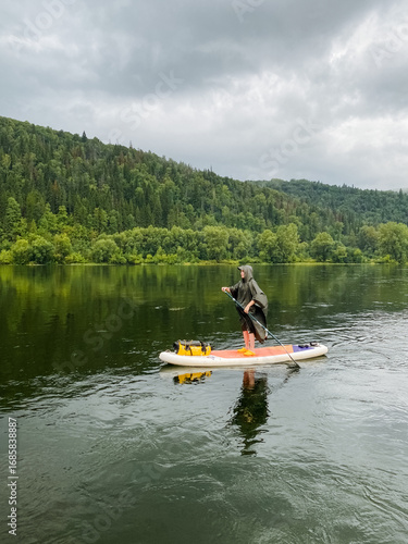 An adventurous individual is paddleboarding on a serene and peaceful lake that is beautifully surrounded by lush green hills, all of which are under a calming, cloudy sky