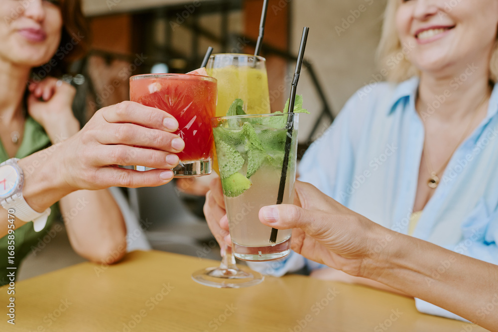 Fototapeta premium Three middle aged Caucasian women clinking glasses with colorful cocktails while smiling at table, hands and partial faces visible, celebrating friendship or special occasion