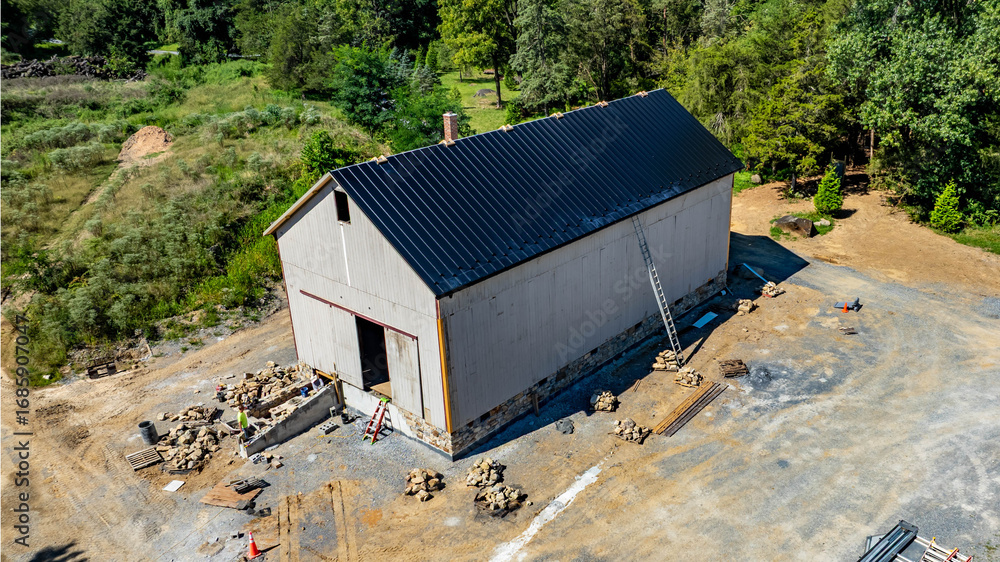 Fototapeta premium Workers are completing the construction of a modern barn in a rural location. The site is organized with materials scattered around and landscaping visible in the background on a sunny day.