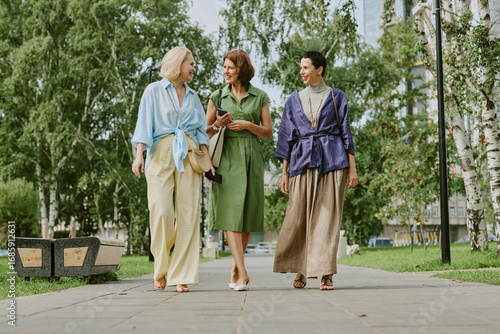 Fotografie Three middle aged women walking together outdoors in park, smiling and talking,