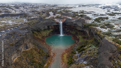 An aerial view of Salto del Agrio waterfall plunging into a turquoise lagoon. The dramatic volcanic landscape is dusted with snow, highlighting a wild and majestic destination
