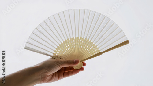 A hand holding a simple white folding fan against a bright white background.