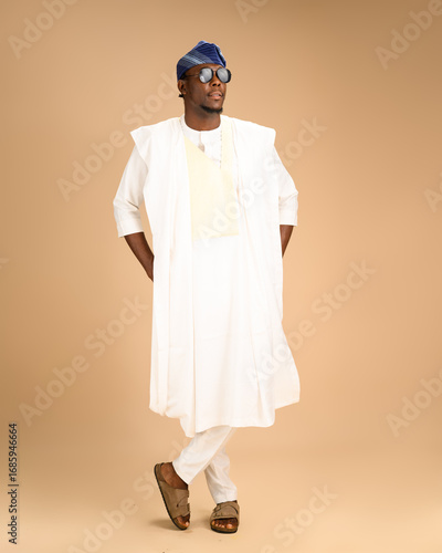 A Nigerian man in full white agbada traditional attire with a blue fila cap and sunglasses poses confidently with crossed legs in a studio setting.