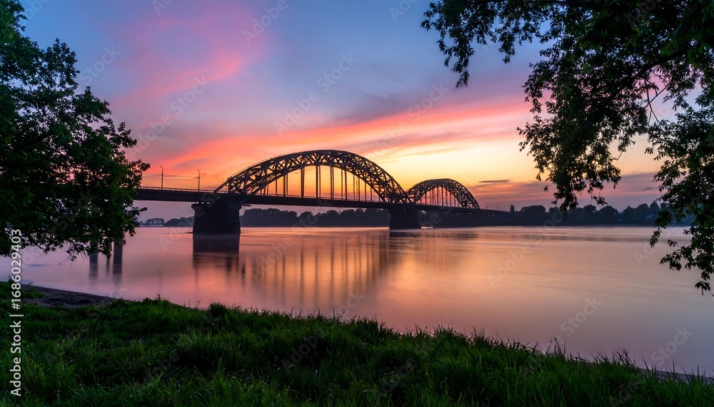 Naklejka premium A serene riverside view at dawn, showcasing a magnificent arched bridge reflecting in the calm water under a vibrant sunrise sky.