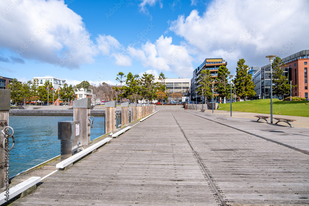 Obraz premium Wooden boardwalk or pedestrian walkway along the waterfront in Geelong, Australia, with modern buildings, seats and open park space. Concept of urban waterfront design, coastal public recreation.