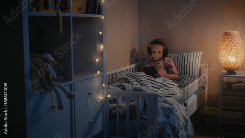 Serious girl, 5-7 years old, sits quietly in bedroom using tablet. Soft string lights create a serene atmosphere during night.
