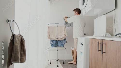 Caucasian man, 35 years old, folds clothes in organized laundry room. Daylight highlights clean towels and drying rack.