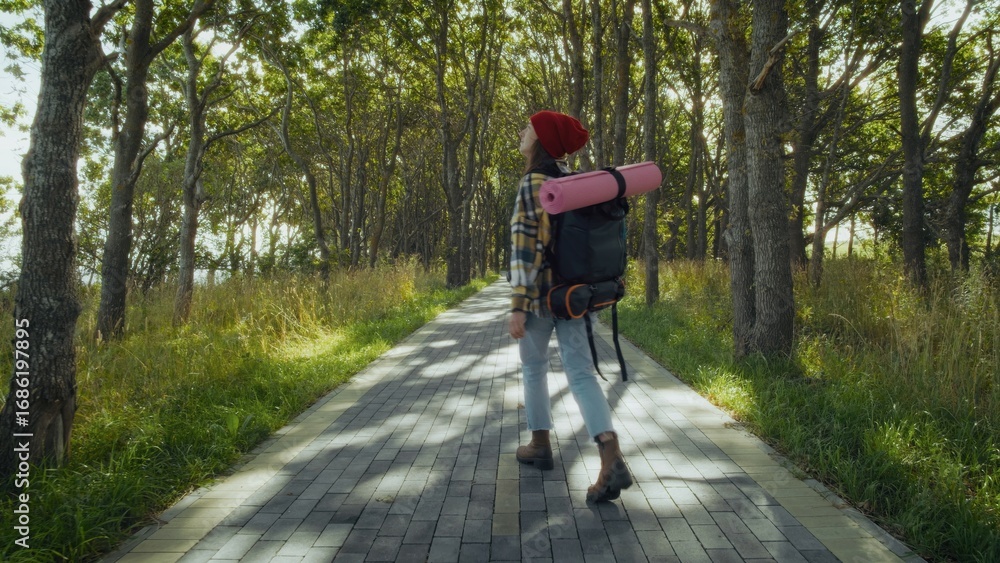 Fototapeta premium Young woman appears content while walking along paved path in serene park. Carrying yoga mat and backpack, she enjoys nature.