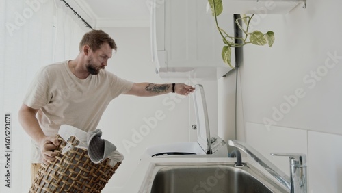 Caucasian man, 35 years old, calmly loads laundry basket into washing machine in bright laundry room. Mundane task during daytime.