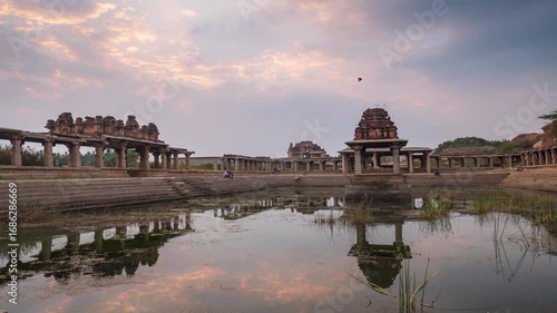 Time lapse: ancient stone temple complex in Hampi, India, reflected in water tank under dramatic evening sky, symbolizing heritage, spirituality, and timeless history