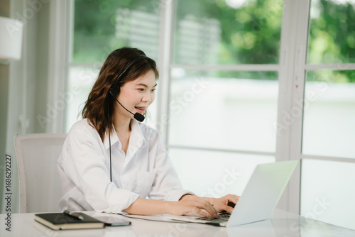 A young woman wearing a headset works at a desk in a call center, smiling as she provides customer support and online assistance.