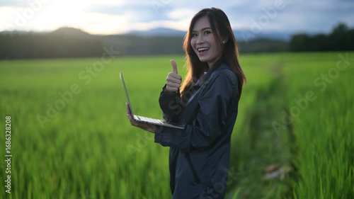 A joyful woman gives a thumbs-up while holding a tablet in a lush green field, showcasing happiness and outdoor engagement.
