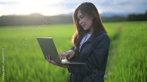 A woman works on a laptop in a lush green rice field under a scenic sky, blending technology with nature.