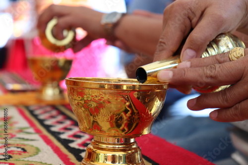 Close-up of hands and cup, Thai Buddhist ritual pouring water from a small golden cup. Hands of a person performing a merit-making ceremony.