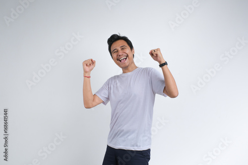 A cheerful young man in a white t-shirt smiling and raising his fists in celebration, expressing happiness and excitement. Studio shot on plain background.